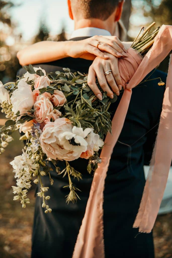 A bride and groom in a close embrace, the bride showing off her bouquet with a peach ribbon