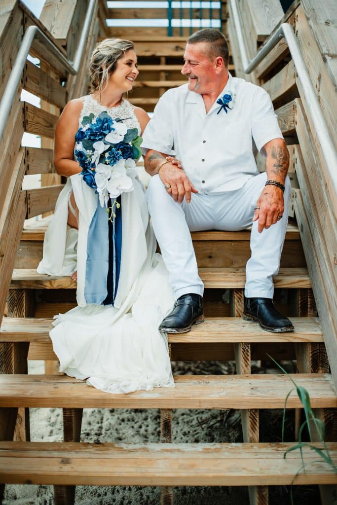 Couple in all white attire, sitting facing each other on a tall wooden staircase outdoors in Orlando, FL on their wedding day