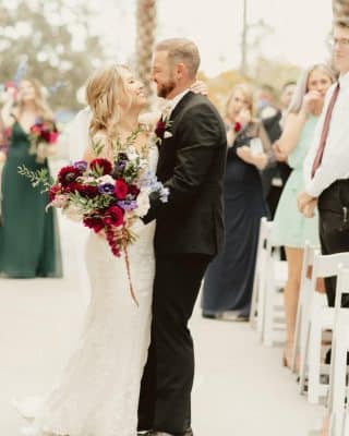 Bride and Groom as they leave their ceremony in an embrace with her red floral bouquet, by Laynie Botanicals