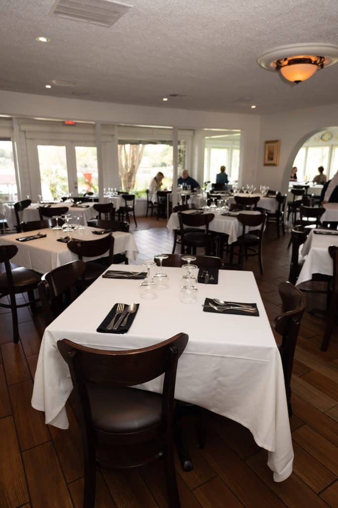 square tables with white table cloths and dark chairs in restaurant with lots of windows