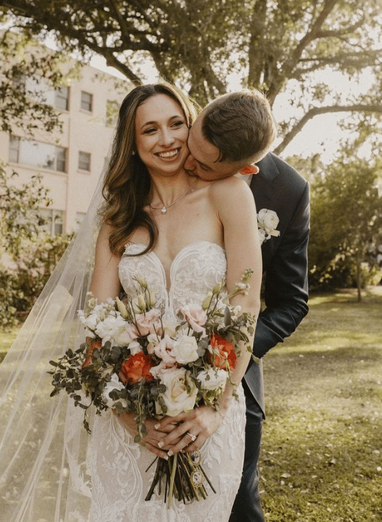 Groom embracing his bride from behind, kissing her neck, bride is holding her bouquet of pink, white and red flowers, Orlando, FL, by Laynie Botanicals