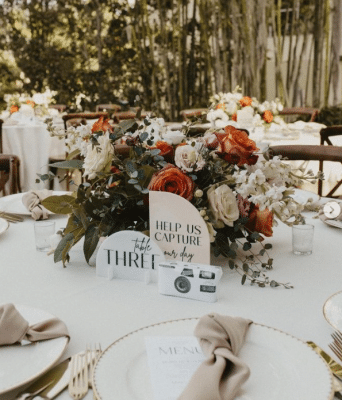 Table at wedding reception with a center floral arrangement, white chargers, tan napkins and a camera to help us capture the moment, Orlando, FL