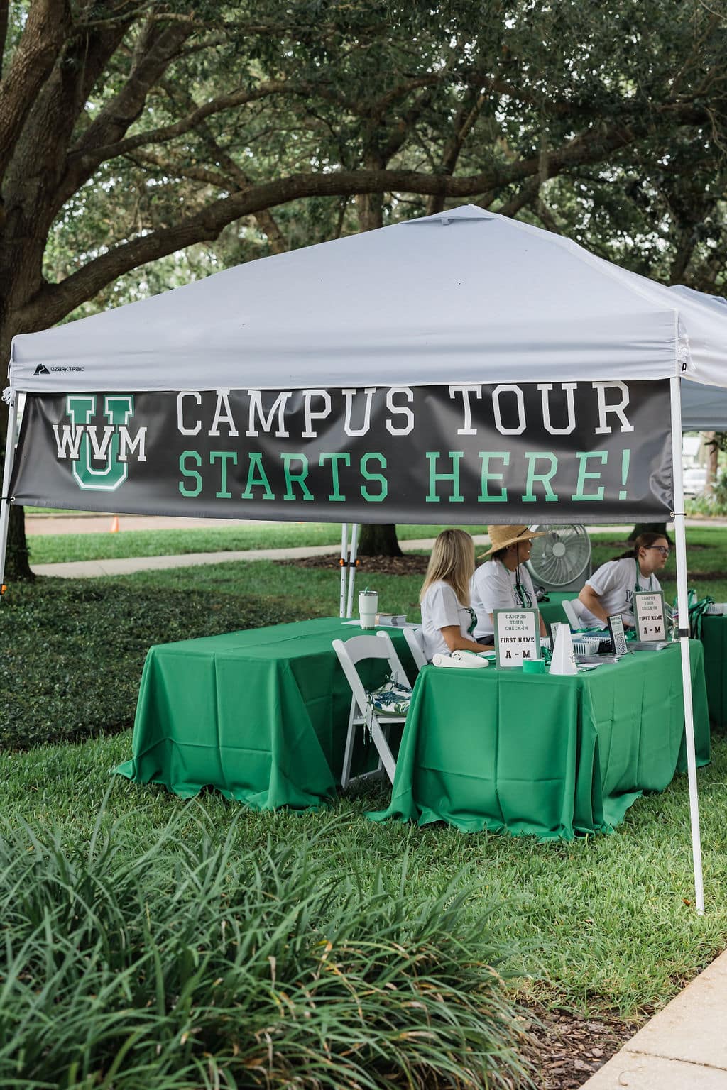 white pop up tent outside in the grass with two tables under it with green linen on it and white chairs between the two tables setup for check in with sign haning from tent