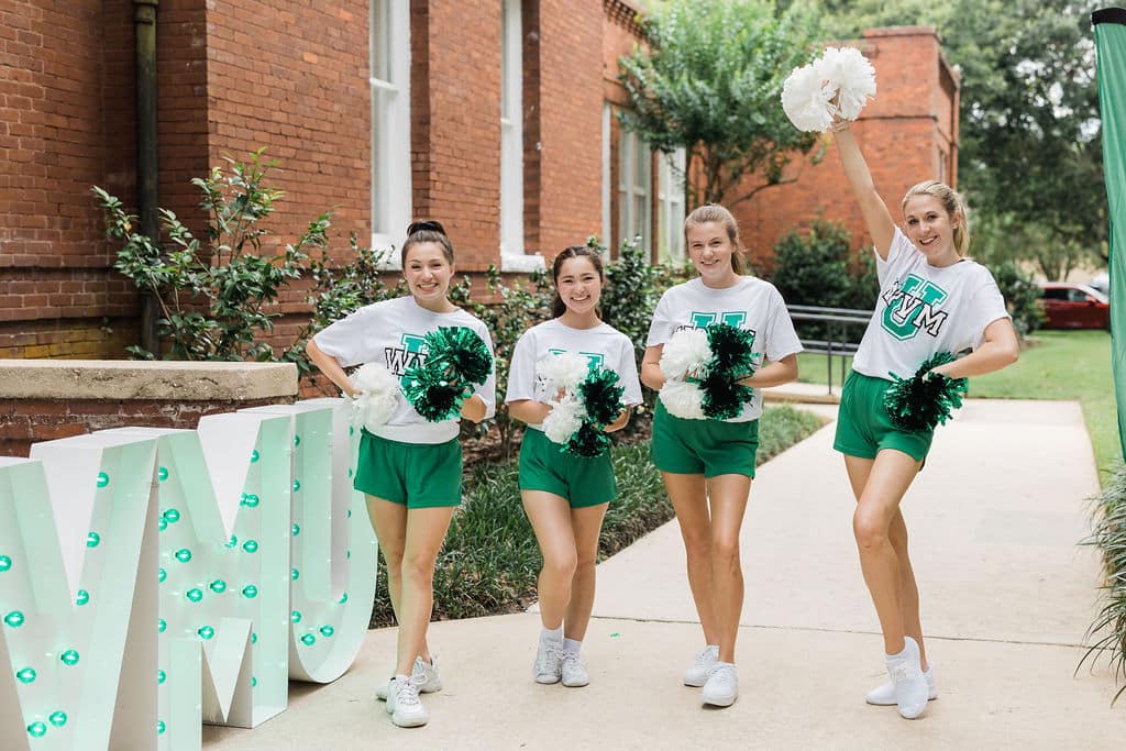 four women in green shorts and white shirts with logos on them holding green and white pom poms on sidewalk in front of brick building