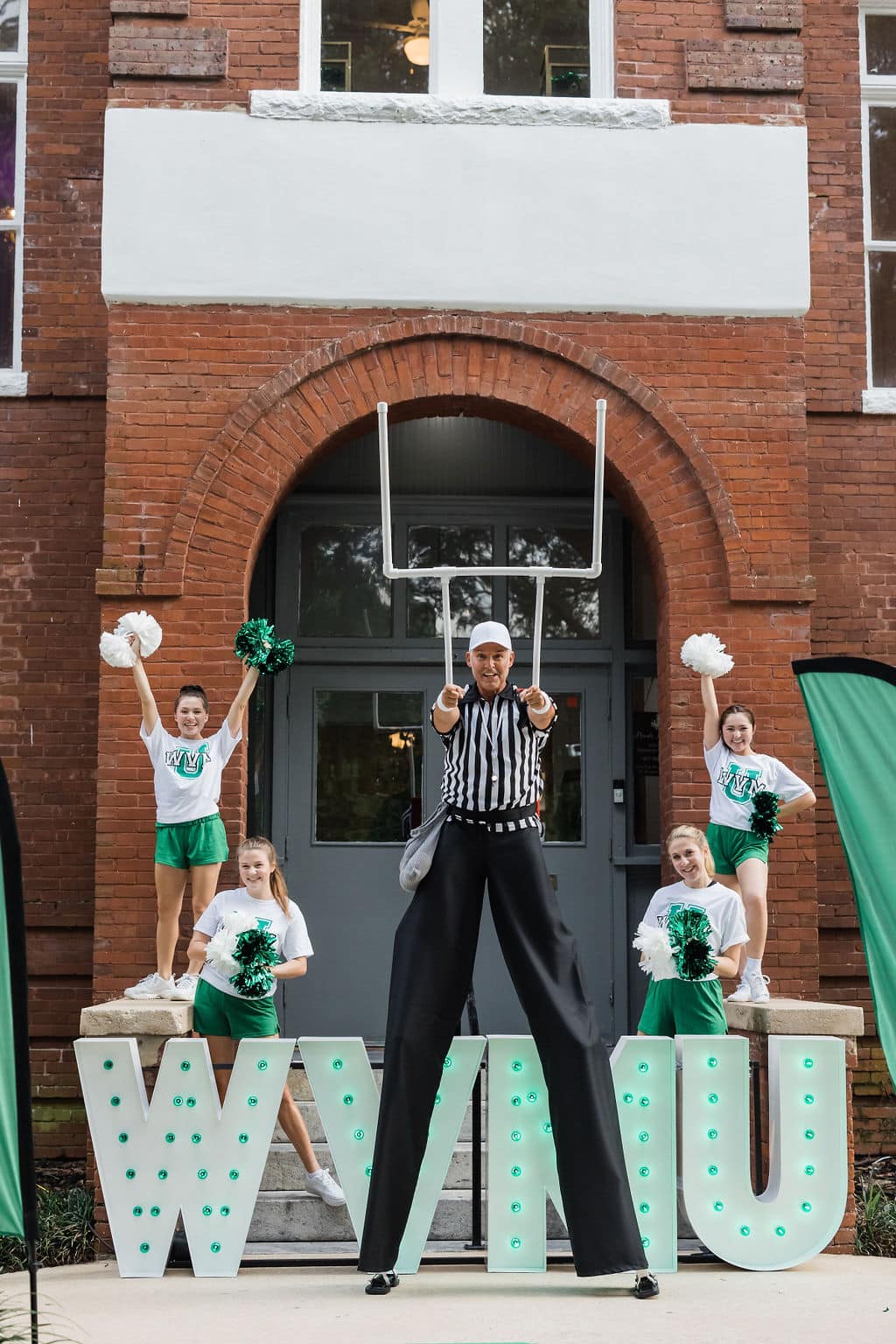 four cheerleaders on the steps of old school house with wvmu marquee letters in front of them and man wearing stilts dresses as a referee in front of them