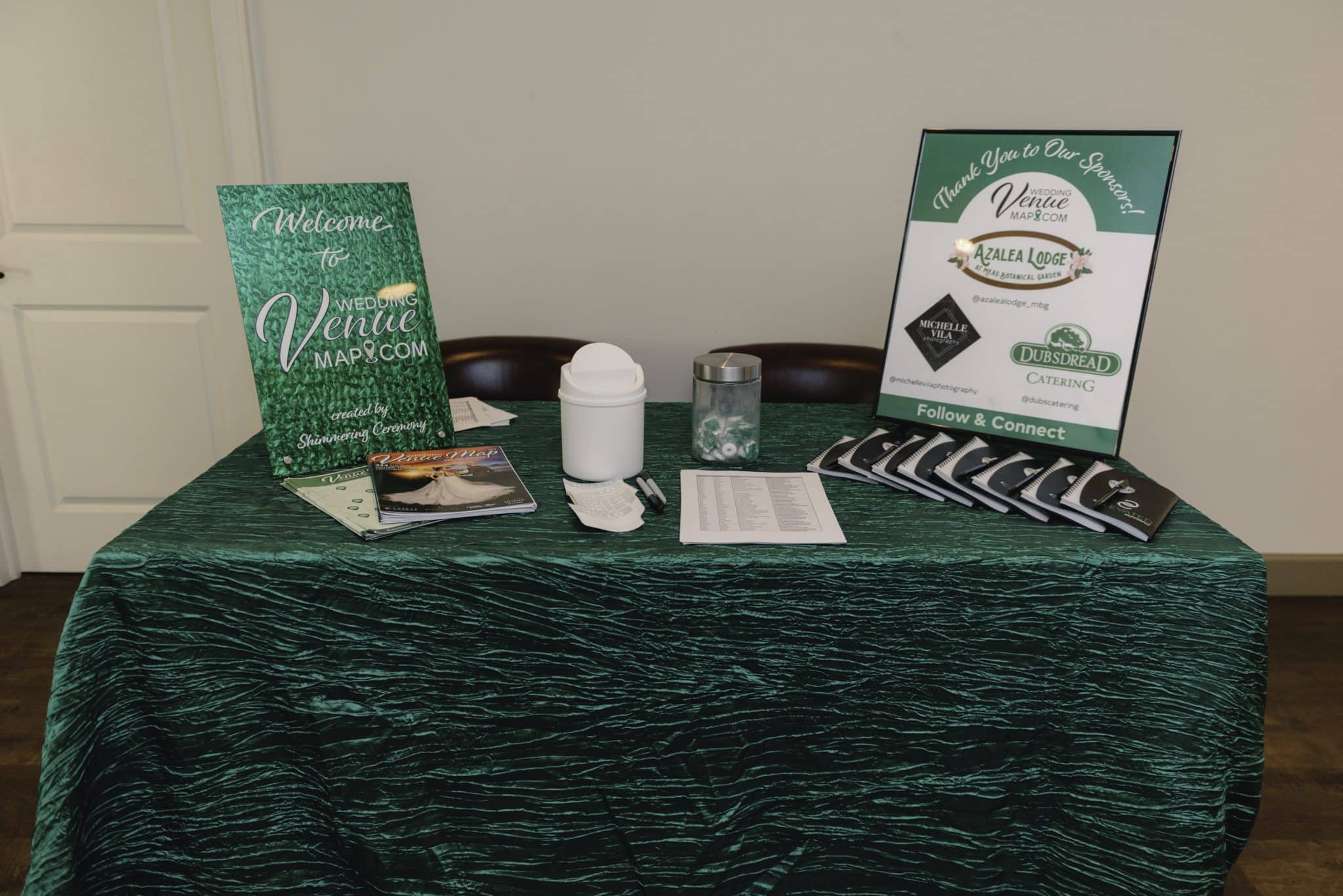 table with green linen on it and two signs standing on top of the table with other registration items like notebooks