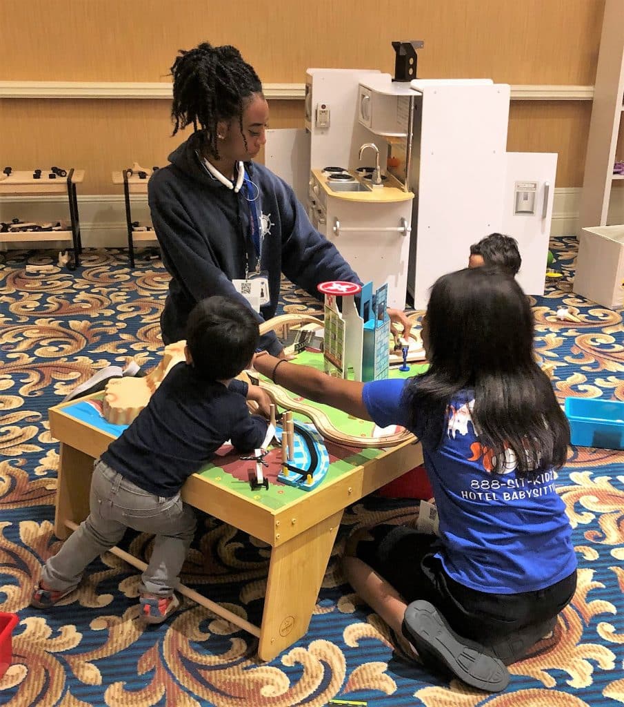 A play kitchen in the background with two staff members and two children using blocks on a mat to build a city, Destination Sitters LLC , Central FL