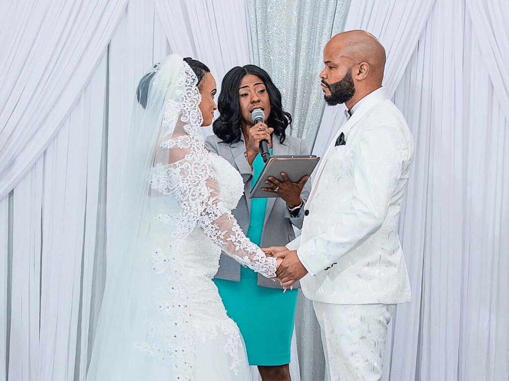 Bride and Groom as they are saying their wedding vows, officiant wearing a teal dress, bride in a lace dress with a lace veil, groom in a white suit, with a white backdrop, Marry Me Now Weddings & Elopements, Central FL