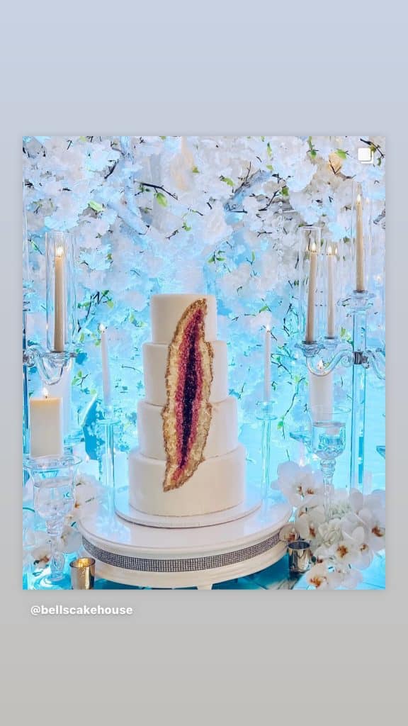 a four tiered white cake, with a cascading design down the side, the cake is on a white stand surrounded by white flowers and lit candles, Central FL