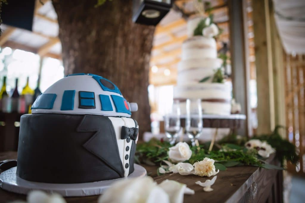 Two cakes, the first in the foreground of R2D2 dressed in a tuxedo, the second, a four tiered white cake on a wooden table with greens as decorations, The Naked Cupcake