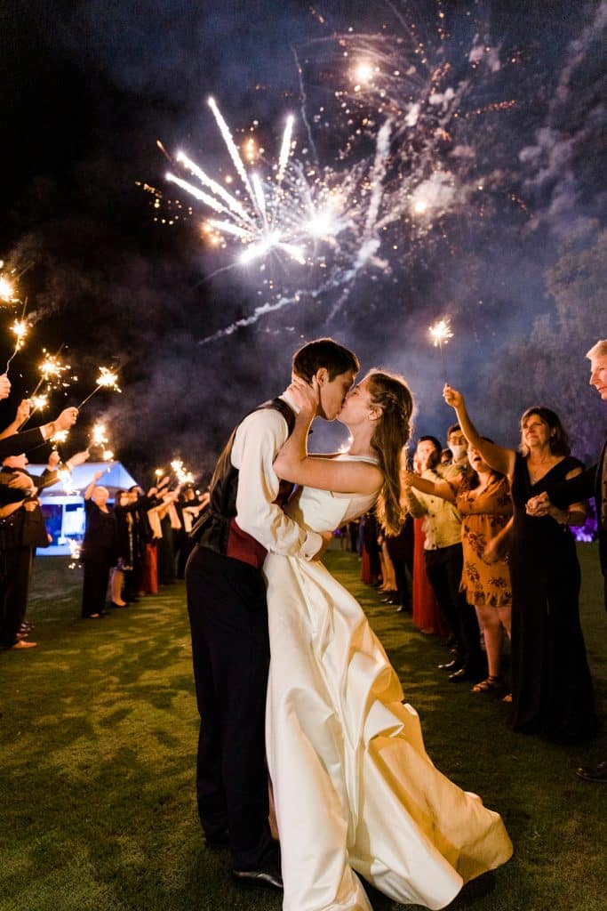 bride and groom kissing in the night sky with fireworks in the background with their guests holding sparklers, Alicia Frost Photography, Central FL
