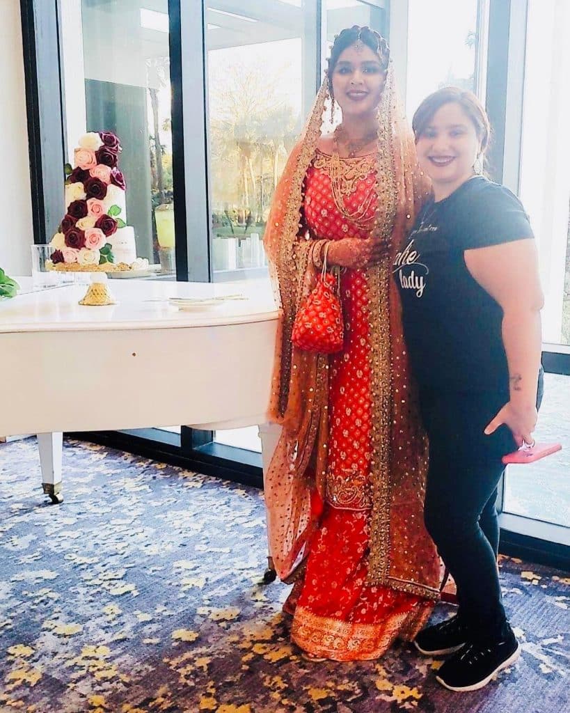 Photo of one of our happy customers with her cake on a table that has pink and red roses on all the tiers, one of our bakers poses with the customer, Central FL