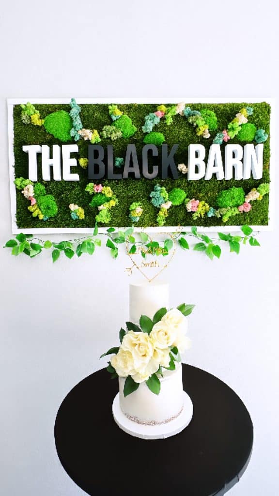 a three tiered cake with white roses and green leaves adorning down the side, sitting on a black table with a sign for the Black Barn behind it, Central FL