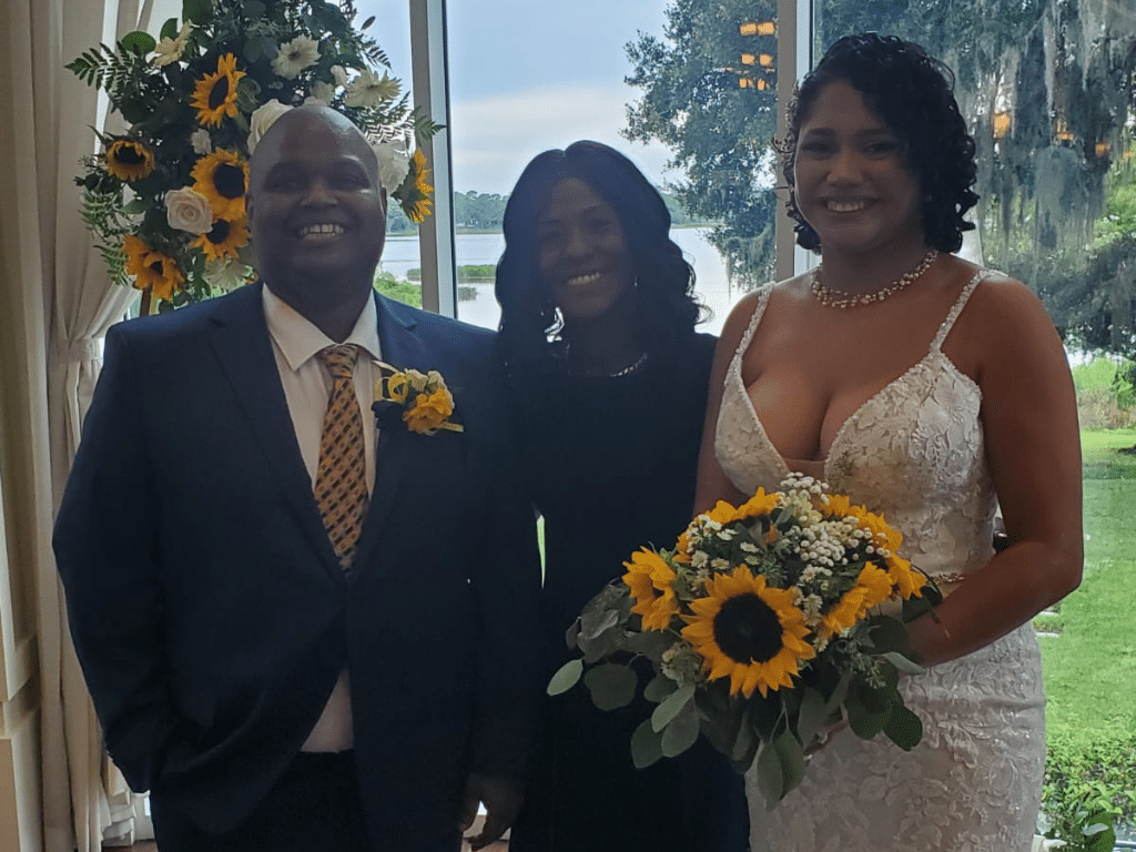 A beautiful couple with their celebrant, bride holding a bouquet of sunflowers, sunflower arrangement in the background, view of the water in the background, Marry Me Now Weddings &B Elopements, Central FL