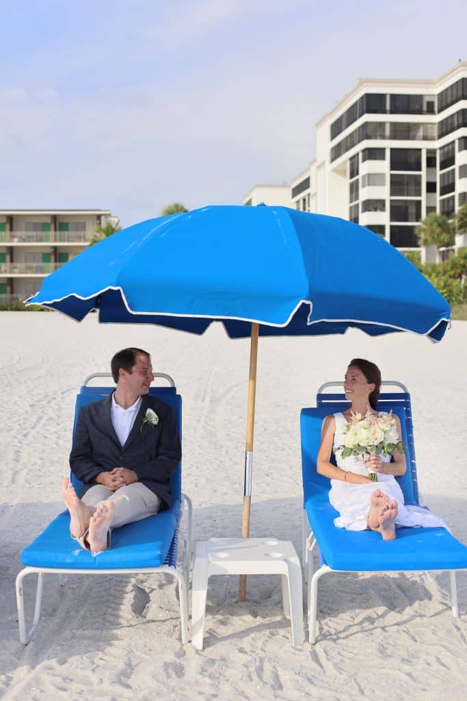 wedding couple sitting in blue lounge chairs under a blue umbrella on the beach, with a high rise hotel in the background, JVK Photography, Central FL