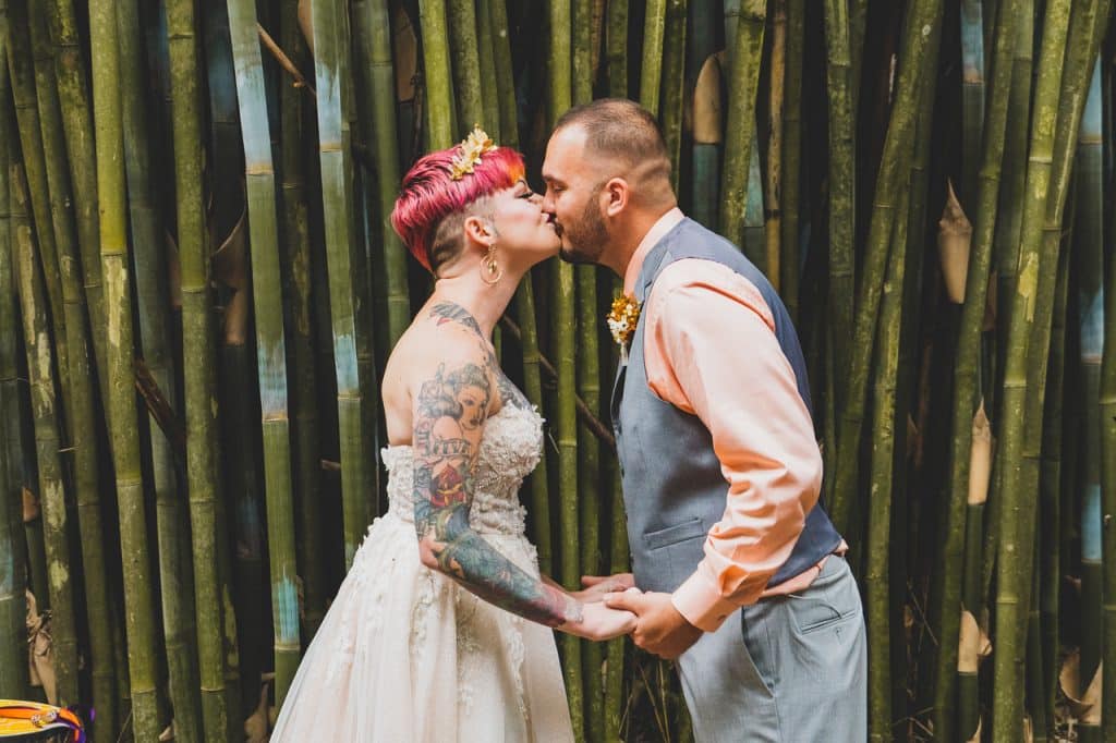 Couple kissing and holding hands, with tall green stalks behind them, bride is wearing a strapless gown, groom wearing a salmon colored shirt with a grey vest and pants, bride has pink hair accented by a tiara, Uptown Selfies, Central FL