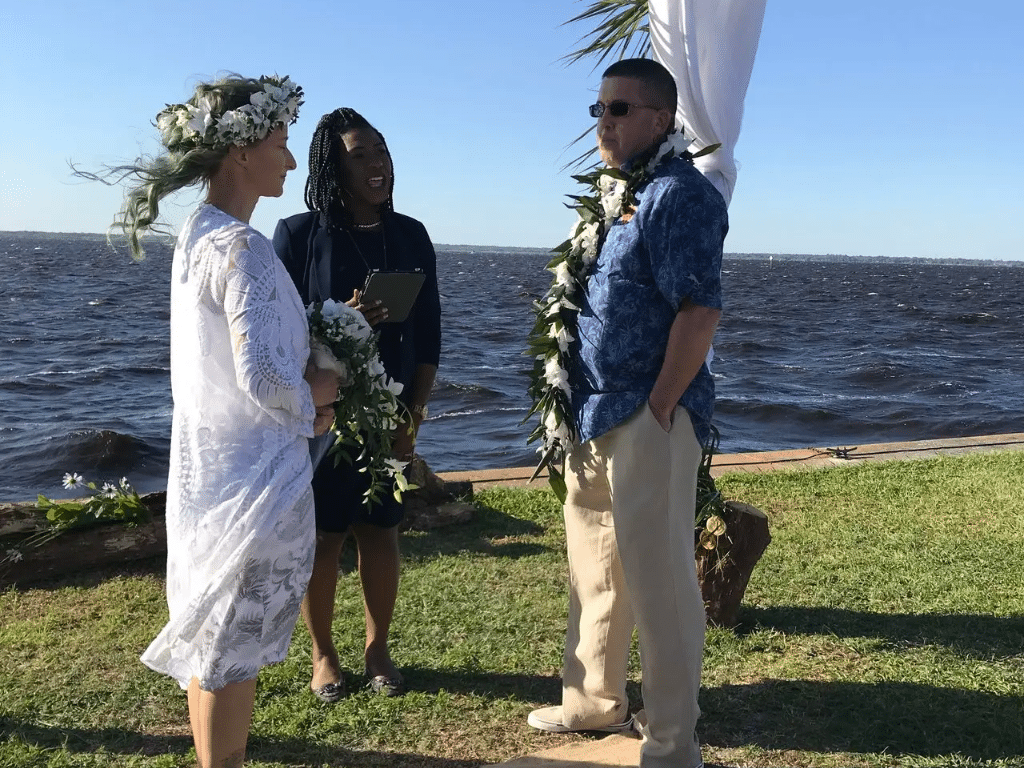 Wedding Ceremony on the beach, water in the background, bride and groom wearing casual attire with flower leis, on their head and around their neck, officiant reading from a book, Central FL