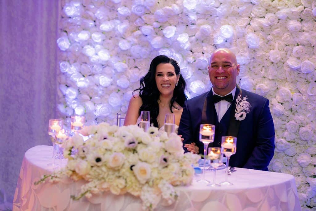Bride and Groom at their head table at their reception, with a white flower backdrop, cascading white flower arrangement on the front of the table with a white tablecloth, Central, FL