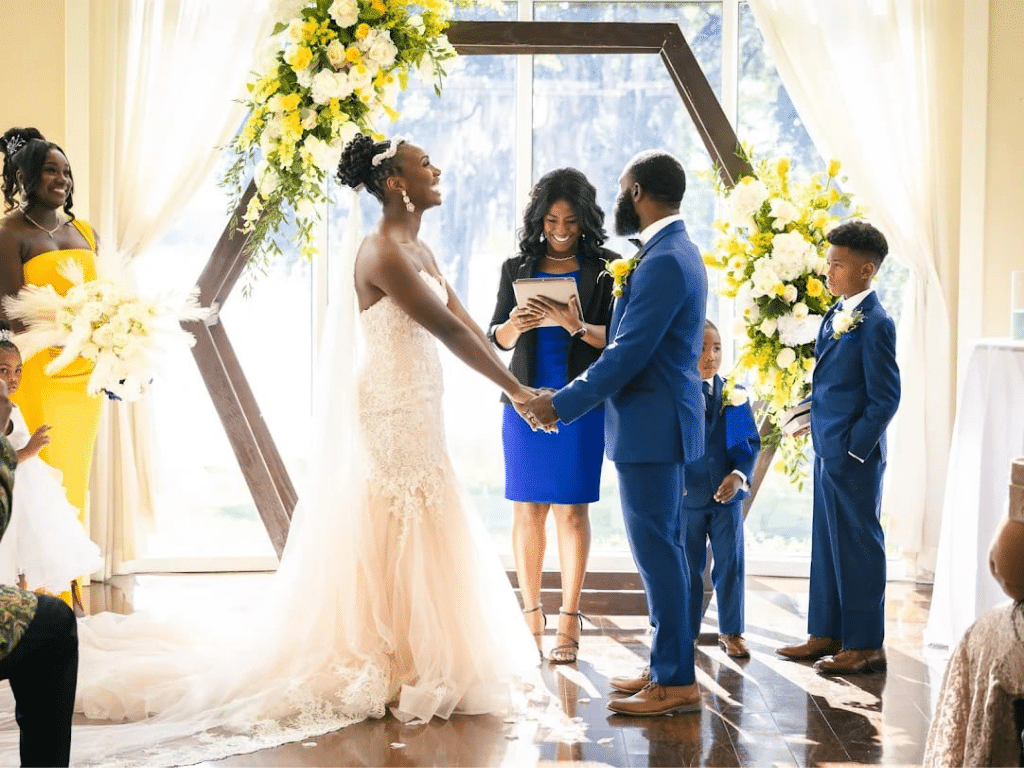 Ceremony at the wooden altar, with a bridesmaid in a yellow dress, a young groomsman in a blue suit, matching the groom, the bride in a light pink gown, officiant wearing a blue dress, Central FL