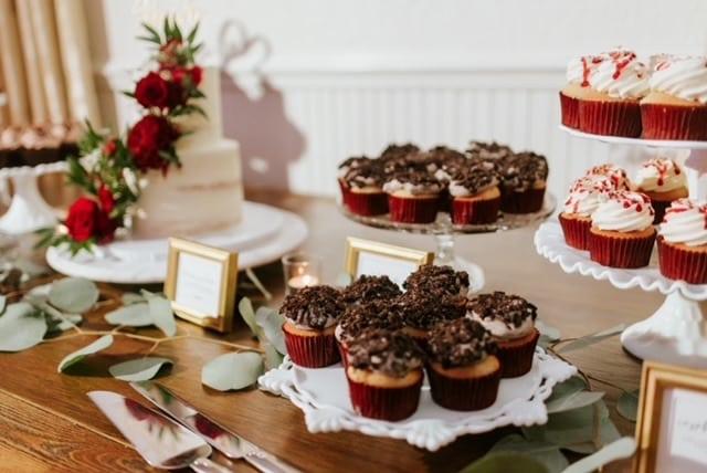 Table display of desserts, including cupcakes on white platters and a two tiered cake with red flowers, and green leaves around it, Central, FL