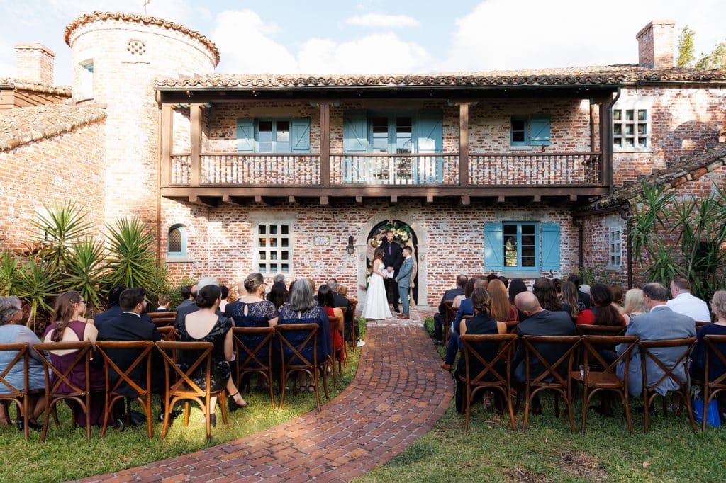 Outdoor wedding ceremony, guests sitting in brown wooden chairs, bride and groom at the front of the aisle, in front of their venue's door, a brick building with blue shudders, Central FL