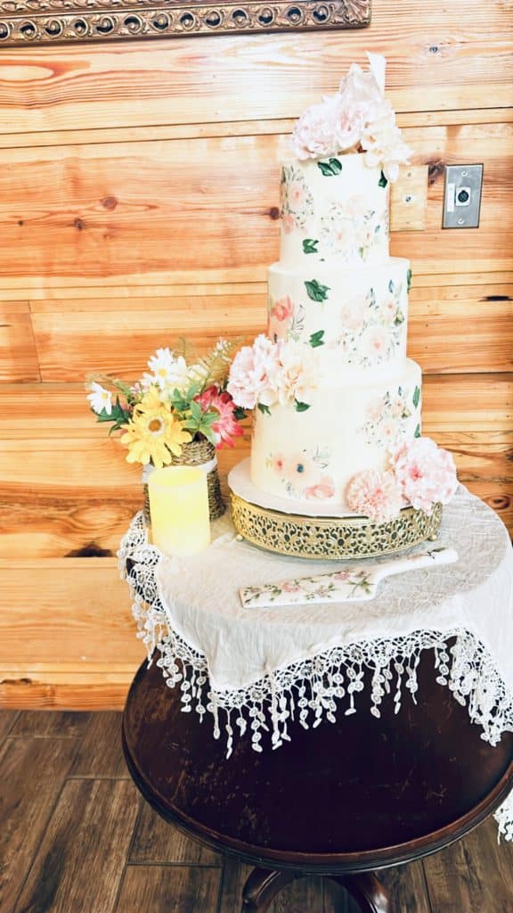 Three tiered cake on a black table with a white fringed tablecloth, cake is sitting on a gold cake stand with an ornamental design, pink flowers on each laters and on top, Bells Cake House, Central FL