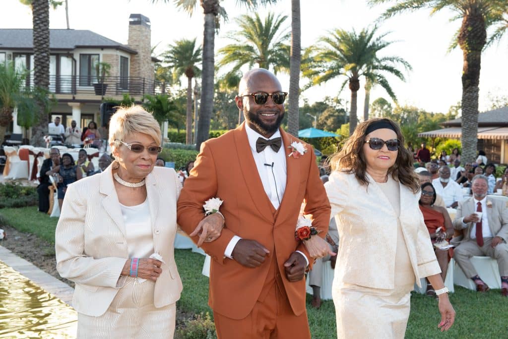 Groom walking down the aisle with his mother and future mother in law, wearing a deep orange suit, outdoors, with palm trees in the background, Central Fl