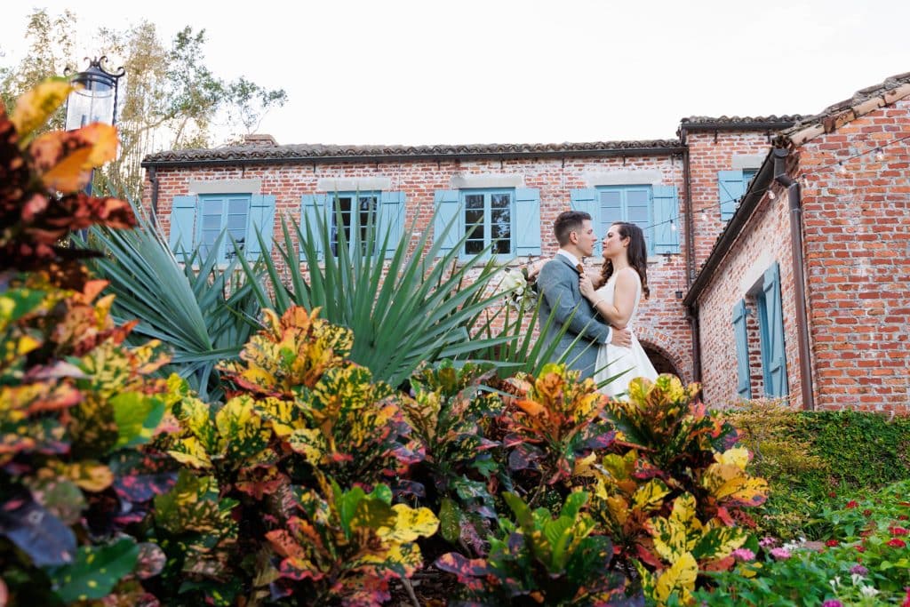 Couple embracing each other, standing in the grass with vibrant flowers in the foreground and a brick building with blue shudders in the background, Central FL