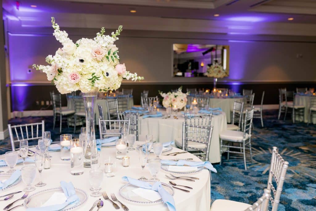Zoomed out view of a ballroom set up a for a reception, white chairs and white tablecloths with tall white flower centerpieces with white chargers and flatware, Central FL
