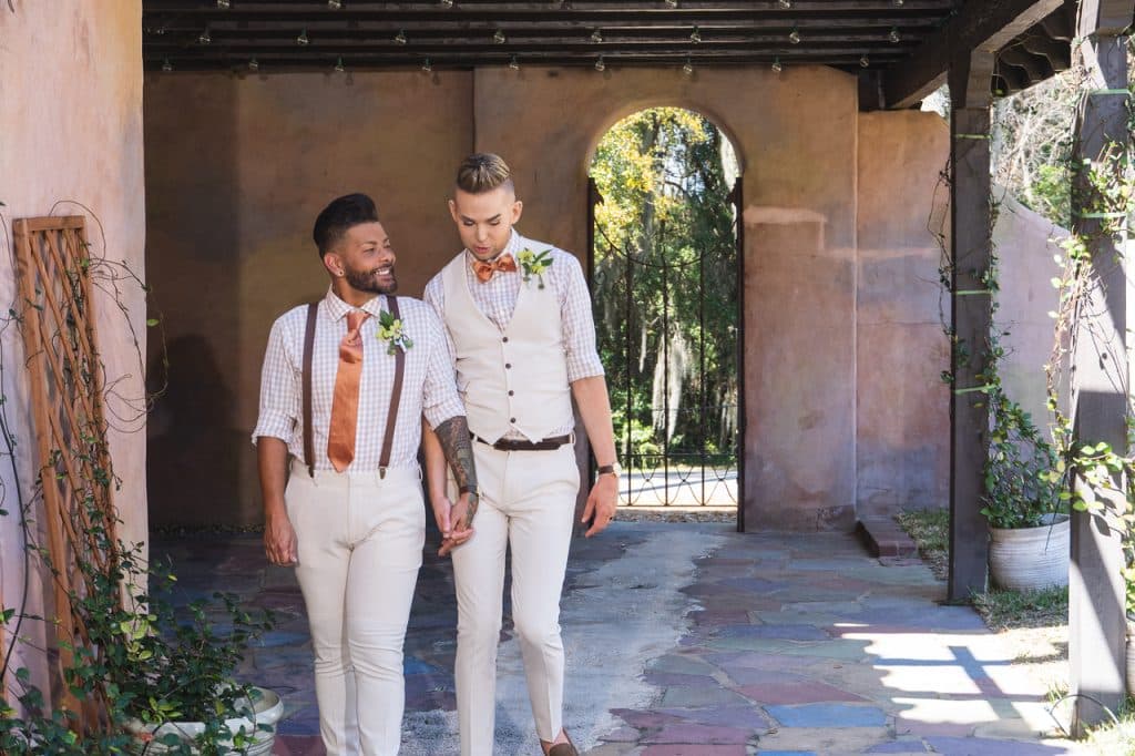 Couple holding hands outdoors at their venue, both in all white suits, one with an orange tie and dark suspenders, the other with a light colored vest and orange bow tie, Central FL