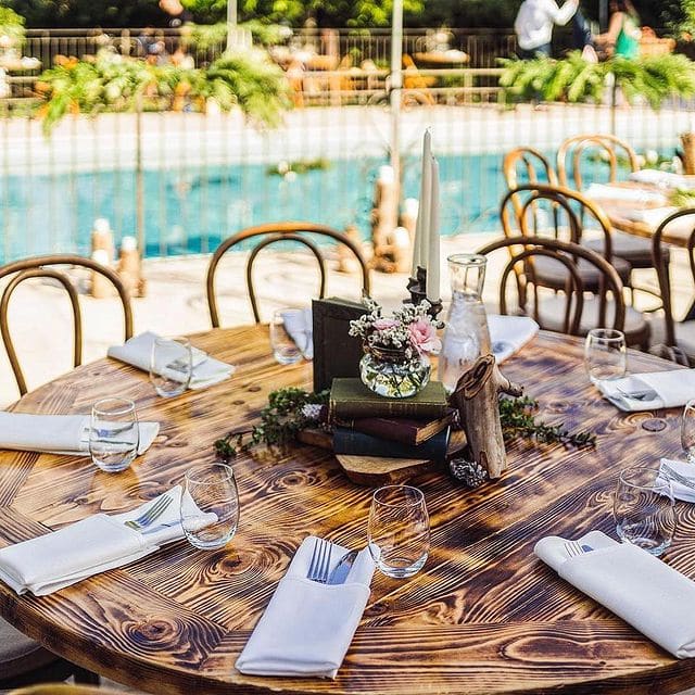 Clear pool in the background of this small event set up with round wooden tables, no tablecloths, white napkins and flatware with books and flowers in the center, Casear Event Rentals Orlando, Central FL