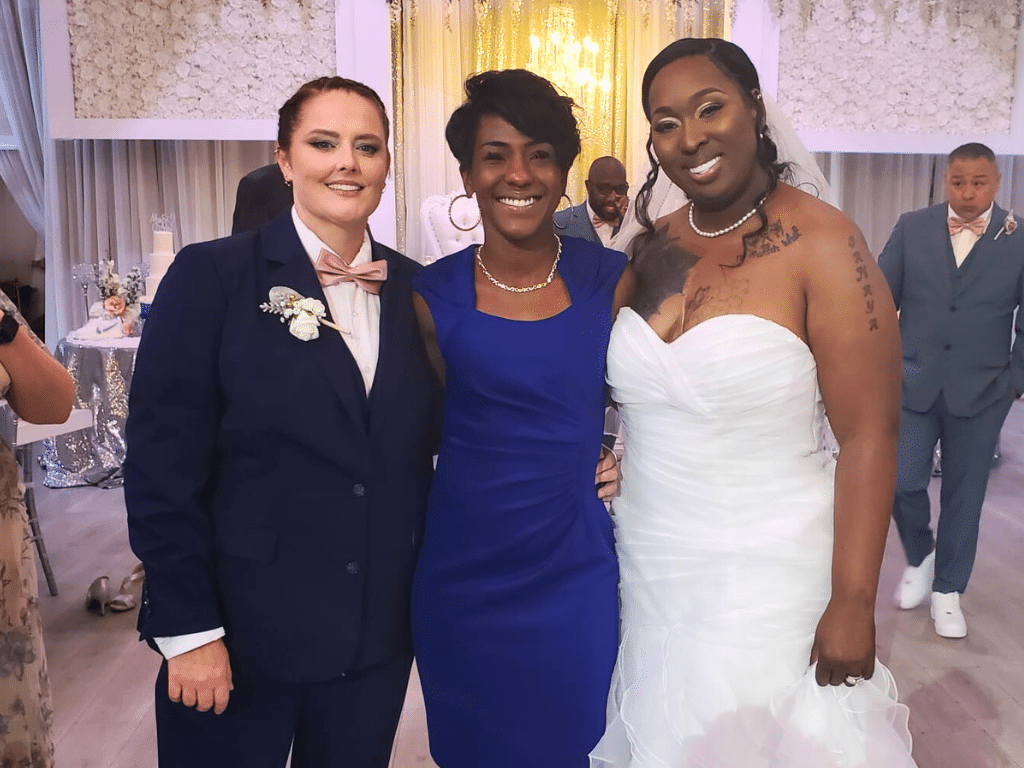 Two women married by their officiant, first woman in a dark blue suit with a pink bowtie, second woman in a white wedding dress, officiant wearing a royal blue dress, Central FL