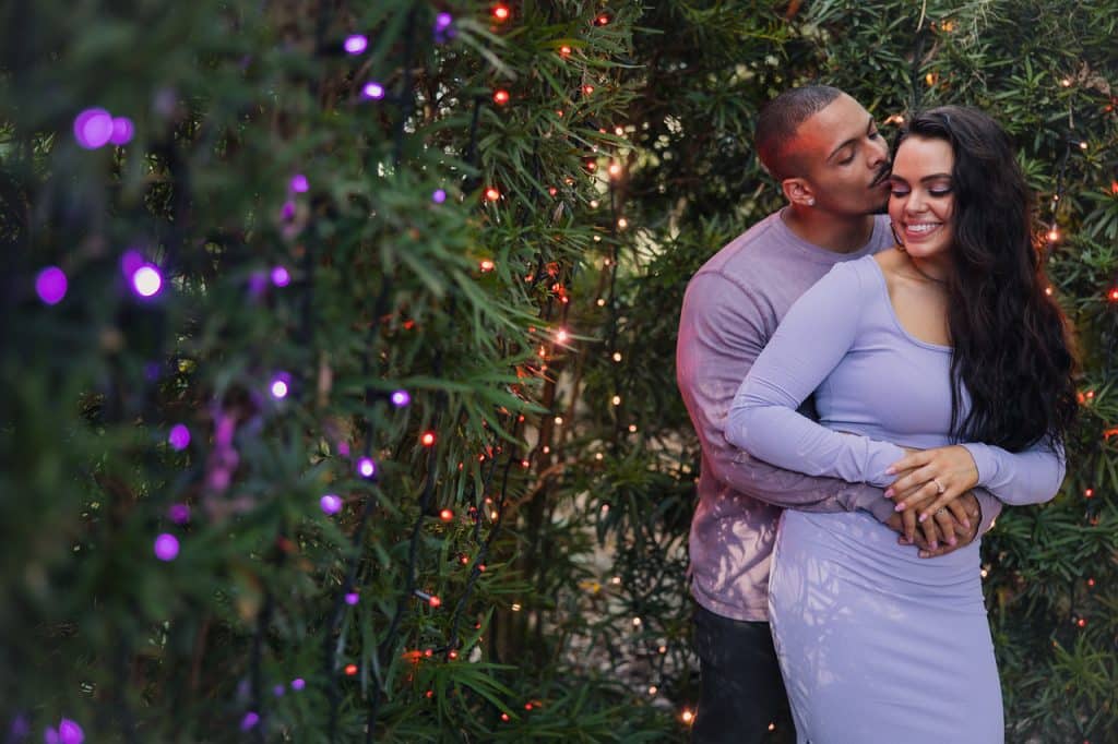 bi-racial couple embracing each other, man kissing her on the cheek, both wearing lavender in an area with pine trees, laced with various colored twinkle lights, Central FL