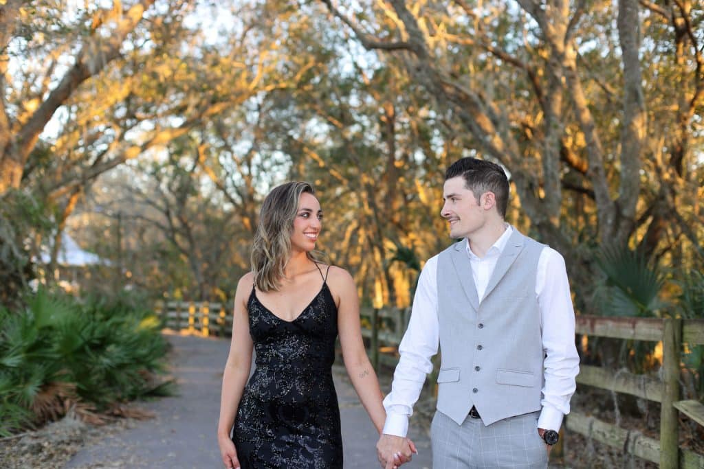 Couple walking and holding hands, on a gravel path with a wooden fence, under trees, Central FL