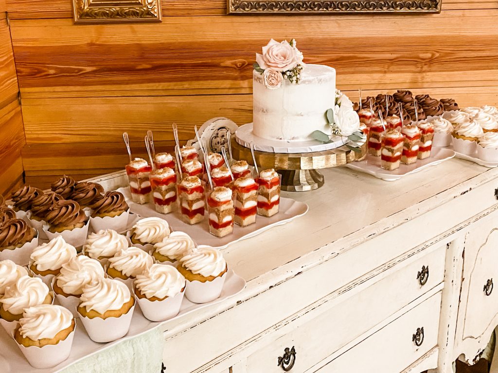 Different view of the white wash dresser of dessert shooters and cupcakes accompanied with a one tiered cake with pink flowers, Central, FL