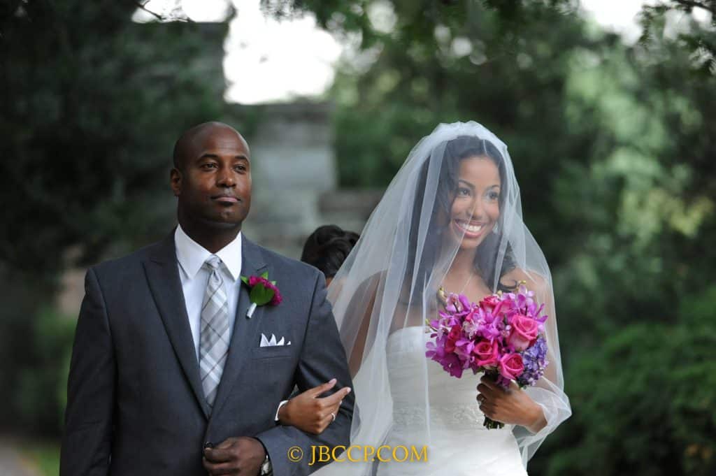 Father walking his daughter down the aisle outdoors, bride carrying a bright pink and purple bouquet