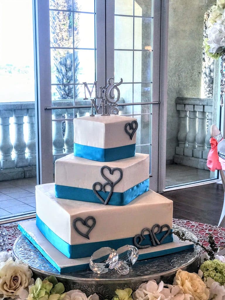 3 later square cake on a silver cake stand, with blue ribbons on each layer, hearts on each layer and Mr & Mrs cake topper on top, large silver rings on the cake stand to represent the engagement ring and wedding bands, Special Treats by Tanya, Orlando, FL