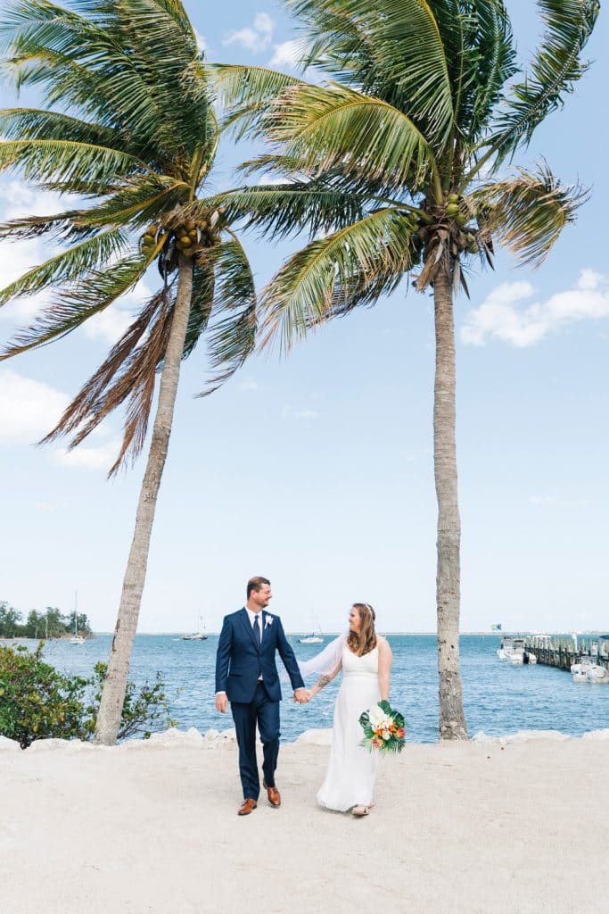 wedding couple holding hands on the beach with the water in background, two palm trees blowing in the breeze, blue skies, groom in a blue suit, bride in her white gown, Alicia Frost Photography, Central FL