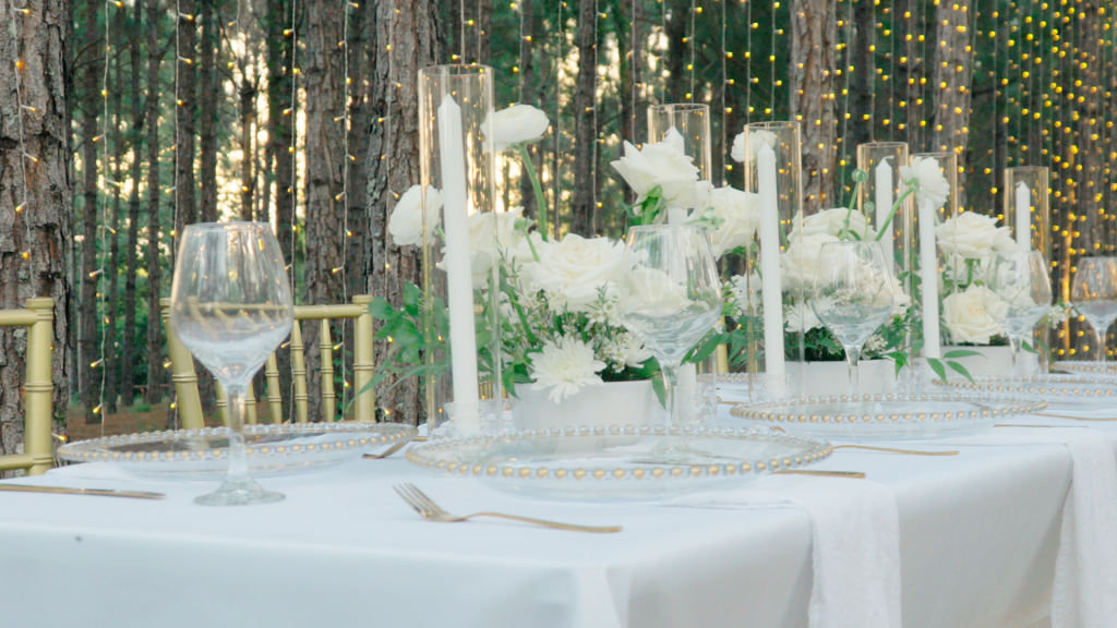 Table set up with white tablecloth, tall white candles, clear glass ware, white flower centerpieces down the center of the rectangular table, trees with handing string lights in the backdrop, Central FL