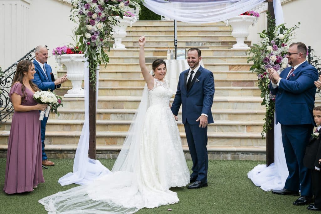 Bride and Groom after their ceremony, in front of a large staircase, wedding attendants on both sides, large flower arrangements on an alter with white sheer material, Central FL