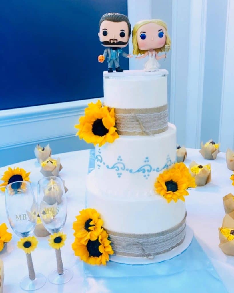 three tiered white cake with sunflowers on each layer, with silver ribbon on the top and bottom layers, cake toppers are figures of a bride and groom, sunflower themed cupcakes also on the table, Central FL