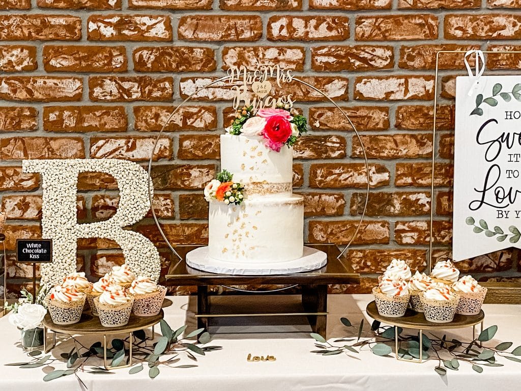 Two tiered wedding cake with red flowers with cupcakes in the foreground, exposed brick backdrop and the letter B on the table, Central, FL, The Naked Cupcake