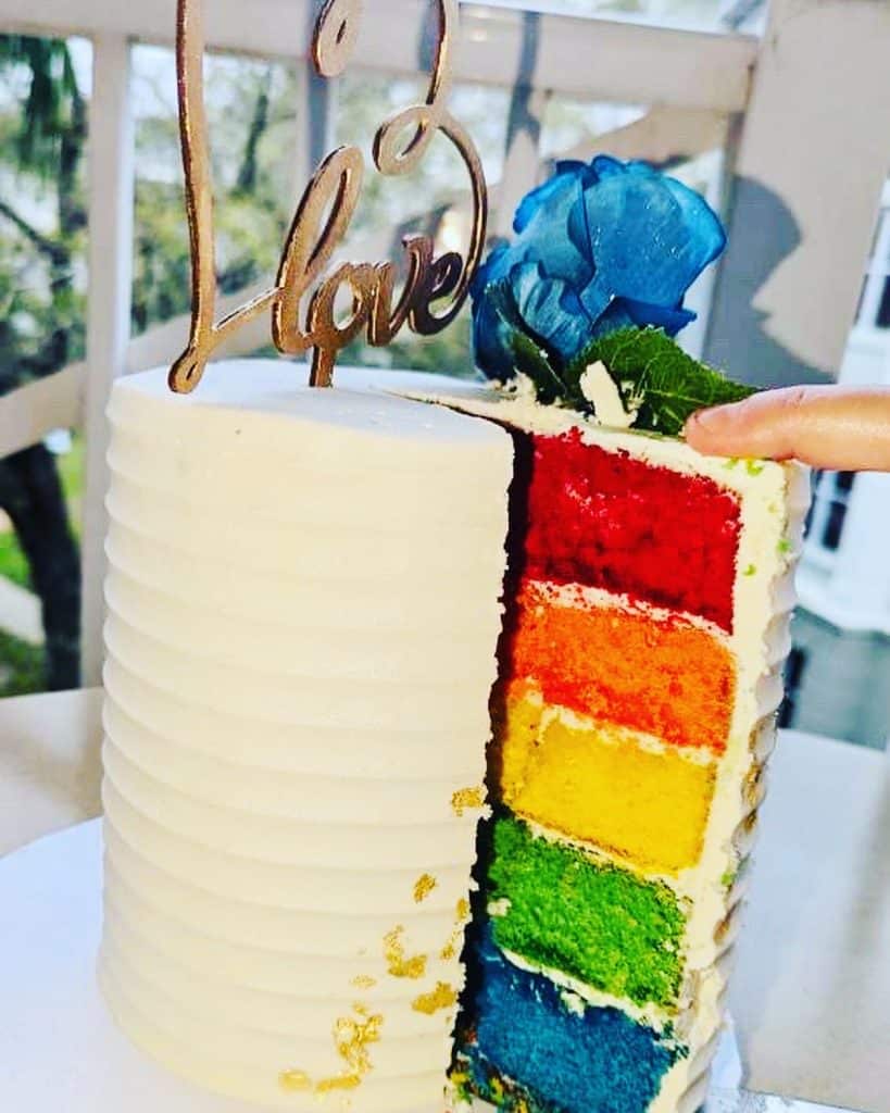 Rainbow cake with a slice missing to show the colors of the rainbow, topped with a cake topper that is a heart and says 