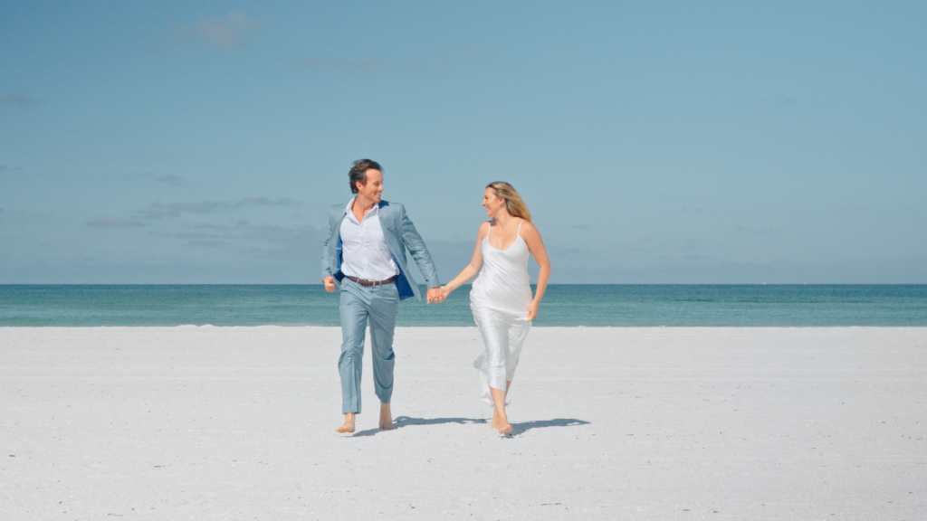 Couple walking on the beach, away from the water, holding hands, barefoot, with a blue sky, BR Focus Films, Central FL