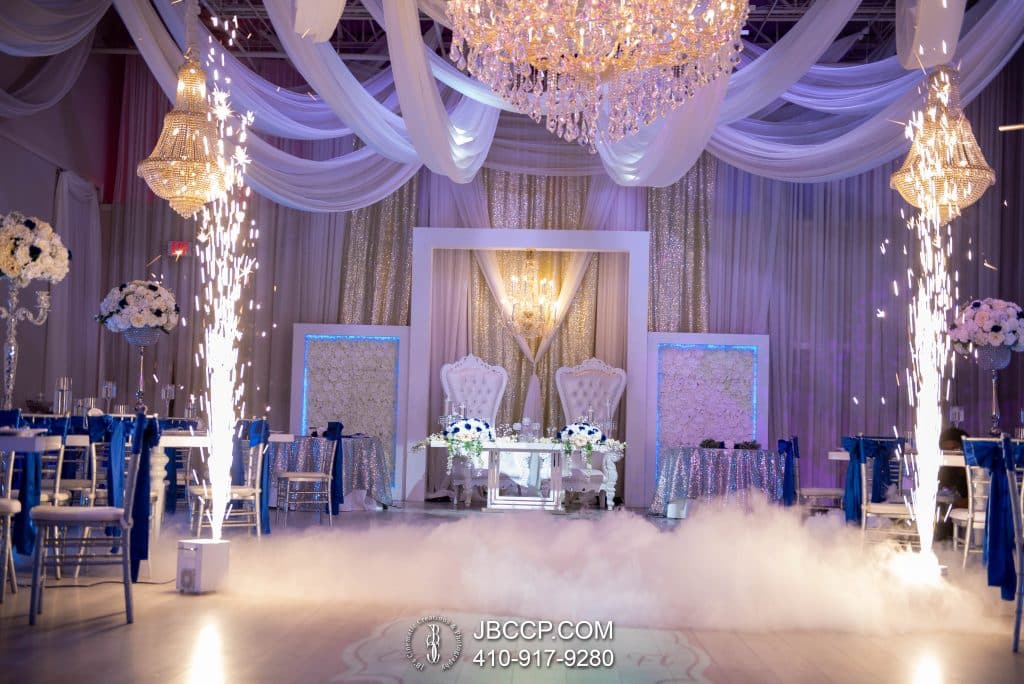 Wedding reception set up, sparklers on the sides, flowing material hanging from the ceiling, uplighting in purple, head table with white chairs and smoke across the floor, JB Cinematic Creations and Photography