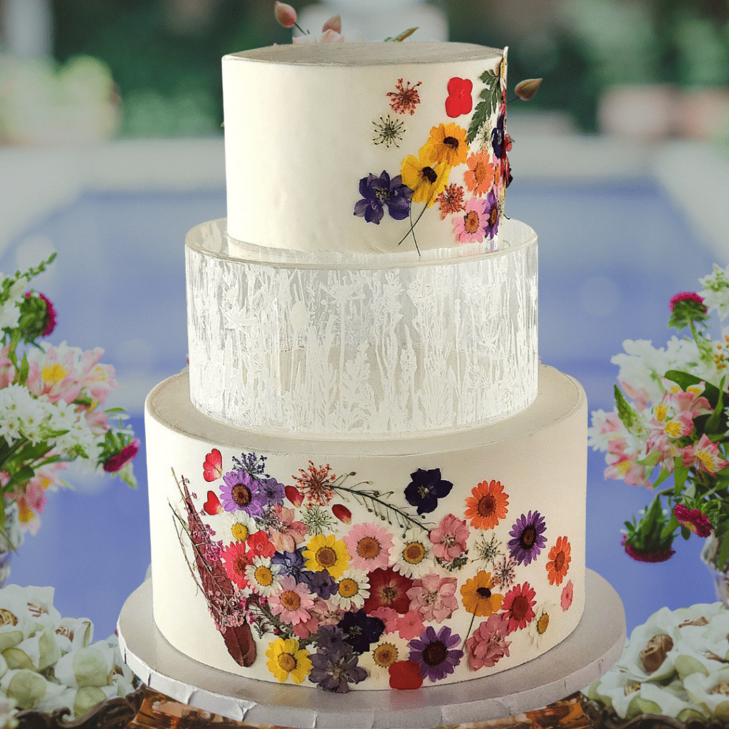 three tiered cake with white fondant, adorned with a variety of flowers on the top and bottom layers, set on a table with flower arrangements on the left and right of the cake, Special Treats by Tanya, Orlando, fl