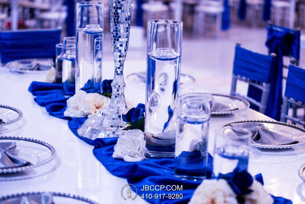 Tablescape of glassware with a blue table runner, clear chargers, blue napkins and silver chairs with blue back coverings, JB's Cinematic Creations and Photography