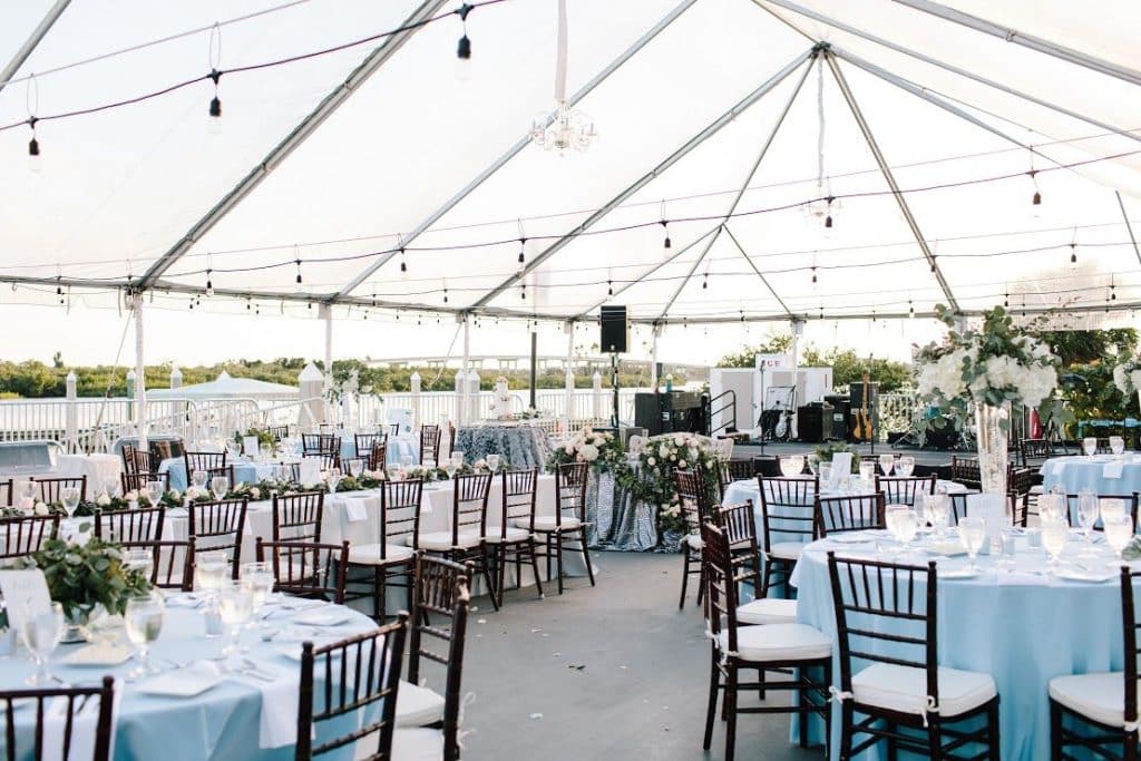 Outdoor tent with string lights hanging from above, tables, with light blue tablecloths, glassware and a view of the water, Central FL