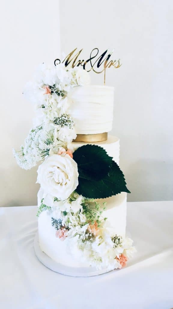 three tiered cake on a white tablecloth, each later has flowers cascading down the side, large green leaf on the middle layer, cake topper is 