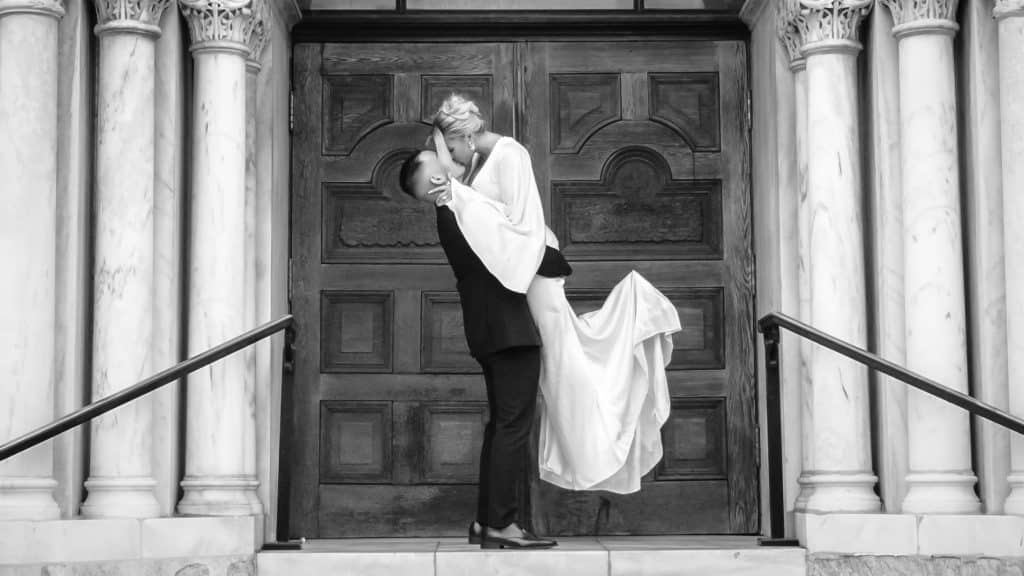 Black and White photo of a groom lifting his bride into an embrace with a kiss, in front of the doors of their venue, BR Focus Films, Central FL